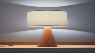 Escena con una lámpara de mesa encendida sobre una superficie clara, con la luz abriéndose en abanico sobre la pared del fondo.