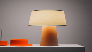 Escena con una lámpara de mesa de base cónica de madera y pantalla color crema, encendida, junto a dos objetos naranjas apoyados a su izquierda.